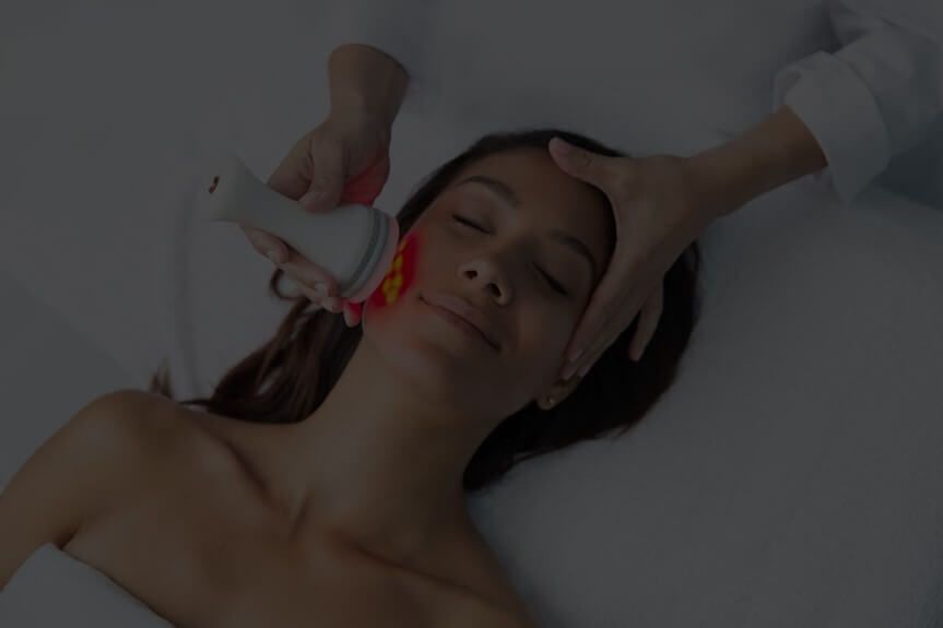 Person lying down with eyes closed, receiving a facial treatment with an LED device.